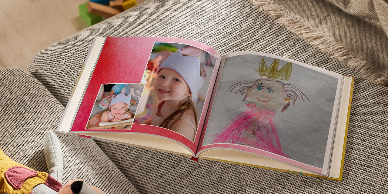 Auf einem Sofa liegt ein geöffnetes CEWE FOTOBUCH neben einem Kuscheltier und einer Decke. Die linke Seite zeigt zwei Fotos von einem lächelnden Mädchen, das eine Krone trägt. Auf der rechten Seite ist eine Kinderzeichnung von einer Prinzessin mit einer Krone zu erkennen. Der Hintergrund der Doppelseite ist mit einem roten Aquarellmuster gestaltet.